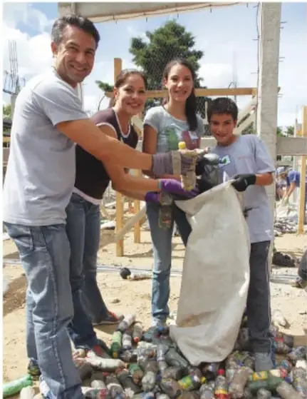 Cuatro personas (un adulto, una mujer, una niña y un niño) recolectan botellas de plástico en un costal, en un espacio al aire libre.