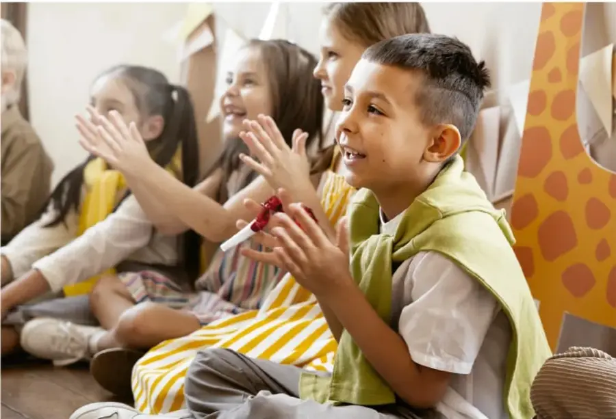 Fotografía de niñas y niños sentados en el suelo del salón, aplaudiendo y sonriendo durante una actividad grupal; al fondo se ve una cartulina decorada con manchas de jirafa.