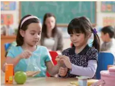 Dos niñas en clase compartiendo materiales; una sostiene una manzana verde.