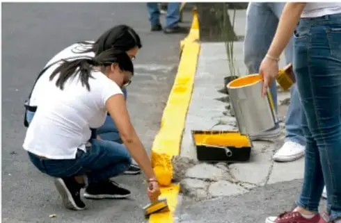 Fotografía de estudiantes pintando de amarillo la línea de una banqueta como parte de un proyecto comunitario.