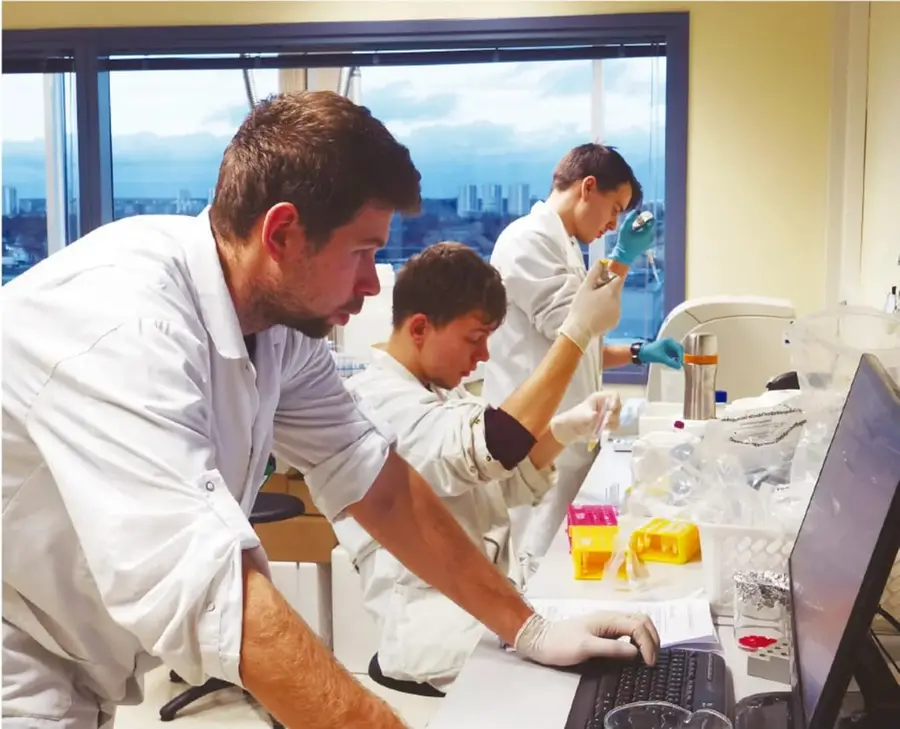 Fotografía de un laboratorio moderno: tres científicos jóvenes con bata blanca y guantes, uno maneja una computadora mientras otros manipulan materiales con punta de pipa; se ve una ventana con rascacielos al fondo.