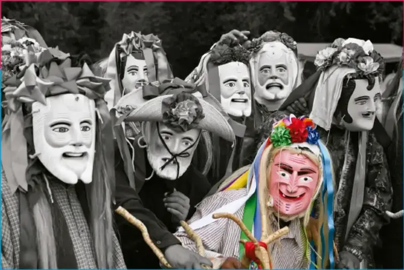 Fotografía en blanco y negro de varios portadores de máscaras tradicionales; una máscara al frente tiene colores (rosa, azul, verde) y cintas de papel.