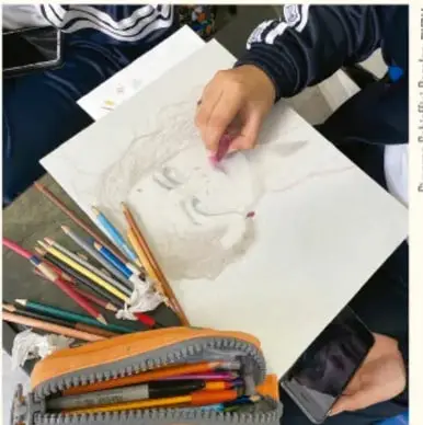 Fotografía de una mano dibujando un rostro a lápiz sobre papel, con colores y una lapicera sobre la mesa.