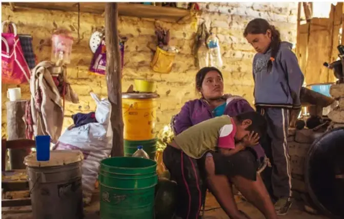 Fotografía de una familia en un espacio rural o marginado, con una mujer y dos niños, en un entorno con paredes de piedra y objetos diversos.