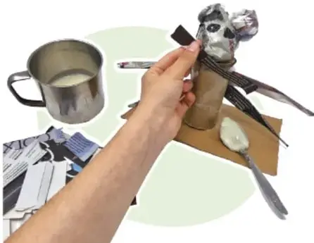 Mano colocando un cuadro de papel sobre el alebrije; a su lado hay una taza metálica con engrudo y cuchara, y trozos de periódico de reúso.