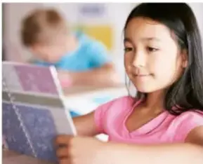 Niña leyendo un libro o cuadernillo en el aula, con otro estudiante al fondo.