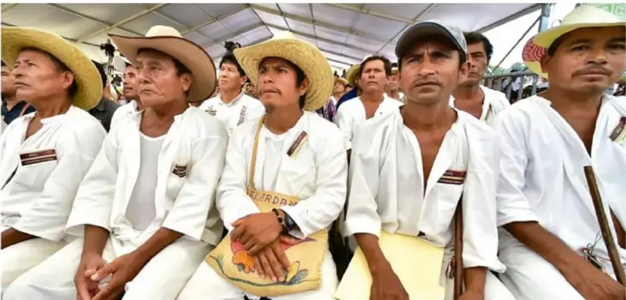 Fotografía de un grupo de hombres indígenas con sombreros de paja y camisa blanca, sentados en un evento comunitario.