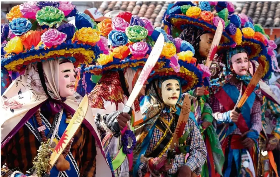 Fotografía de niños y adultos disfrazados en una fiesta tradicional mexicana: sombreros adornados con flores de colores, máscaras, mantas y espadas ornamentales, posando en la calle frente a tejados de teja.