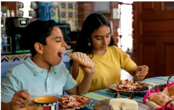 Fotografía de dos niños, un niño y una niña, comiendo en una mesa. El niño sostiene un pan y la niña come de un plato. Están en un ambiente familiar o escolar.