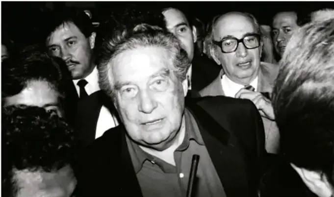 Fotografía en blanco y negro de un grupo de hombres en un encuentro público; en primer plano se observa a un hombre de cabello blanco, posiblemente un escritor o figura literaria vinculada al tema de las posvanguardias.