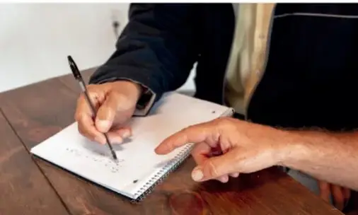 Fotografía de una persona escribiendo a mano en una libreta con espiral sobre una mesa de madera, en alusión a la práctica de escribir crónicas.