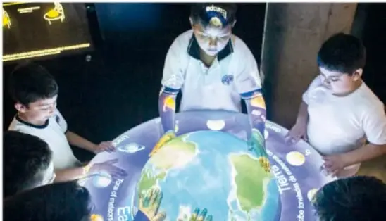 Niños con camisetas blancas alrededor de una maqueta iluminada del planeta Tierra, observando sus continentes.