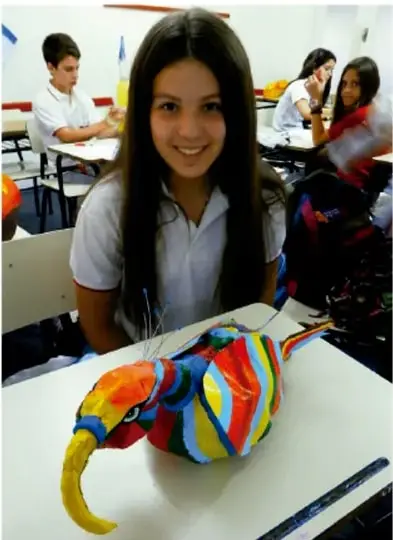 Fotografía de estudiantes en el aula; una niña sonríe con una escultura de animal (tucán) pintada con colores vivos sobre su escritorio.