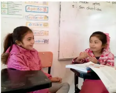 Fotografía de dos niñas de primaria conversando en el aula frente a un pizarrón con meses del año. Crédito: Jesús Eduardo Arteaga Flores, TAMAULIPAS.