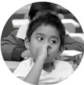 Fotografía circular en blanco y negro de una niña de primaria sentada en el aula, con la mano cerca del rostro, mirando hacia un lado.