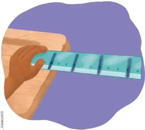 Mano sujetando un plumón azul marcando números sobre una regla translúcida colocada sobre una superficie de madera.