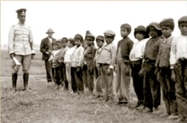 Fotografía histórica en blanco y negro: niños en formación ante un militar, 1915. Archivo Casasola. Muestra a un grupo de niños y adolescentes alineados en un campo, mientras un oficial uniformado los observa.