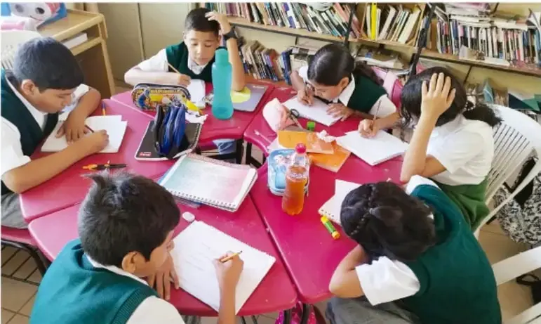 Fotografía de estudiantes de telesecundaria trabajando en equipo alrededor de una mesa rosa, escribiendo en sus cuadernos dentro de una biblioteca aula con estantes de libros.