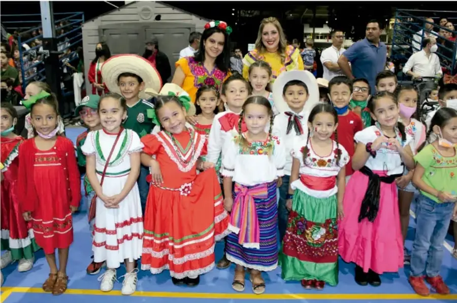 Fotografía a color de un grupo de niños y niñas con vestuarios típicos de distintas regiones, acompañados por adultos, en un espacio amplio con gradas al fondo.