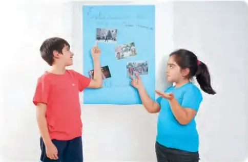 Amanda y Benjamín de pie frente a un papelógrafo azul pegado en la pared; él señala las imágenes y ella explica con las manos.