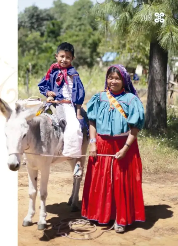 Fotografía de un niño sonriente montado en un burro blanco, con camisa azul y paliacate rojo, acompañado por una mujer con vestimenta tradicional (falda roja, blusa azul, pañuelo morado en la cabeza) que sostiene la soga del burro, en un patio con árboles.