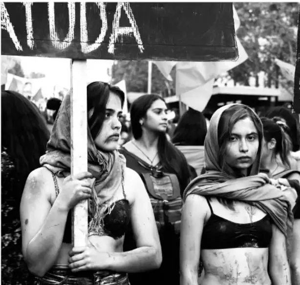 Fotografía en blanco y negro de un grupo de mujeres jóvenes que participan en una marcha. Una de ellas sostiene un cartel con la palabra 'AYUDA' parcialmente visible. Las mujeres portan pañuelos y expresan determinación, lo que ilustra la participación femenina en movimientos sociales.