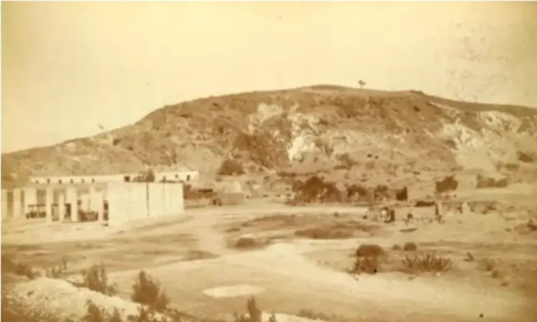 Fotografía antigua en sepia del cerro del Peñón de los Baños, con construcciones y vegetación seca al pie del cerro.