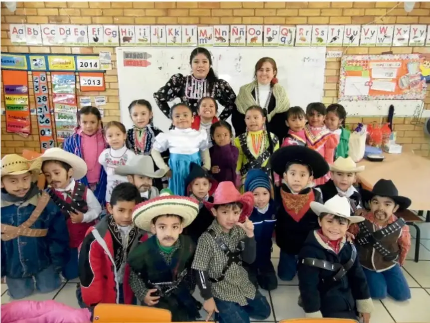Grupo de niños y niñas disfrazados con trajes típicos de México, acompañados por dos maestras, frente a un pizarrón con el abecedario.