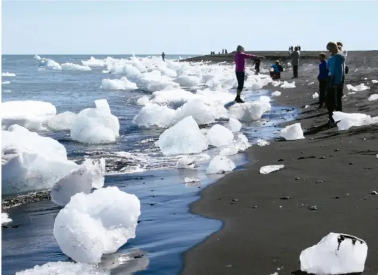 Fotografía de una playa con grandes bloques de hielo blanco arrastrados a la orilla del agua, con varias personas de pie observándolos; evidencia del deshielo asociado al cambio climático.