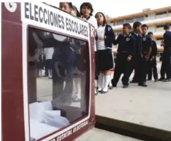 Fotografía de una casilla electoral escolar con la leyenda 'ELECCIONES ESCOLARES' y estudiantes formados.