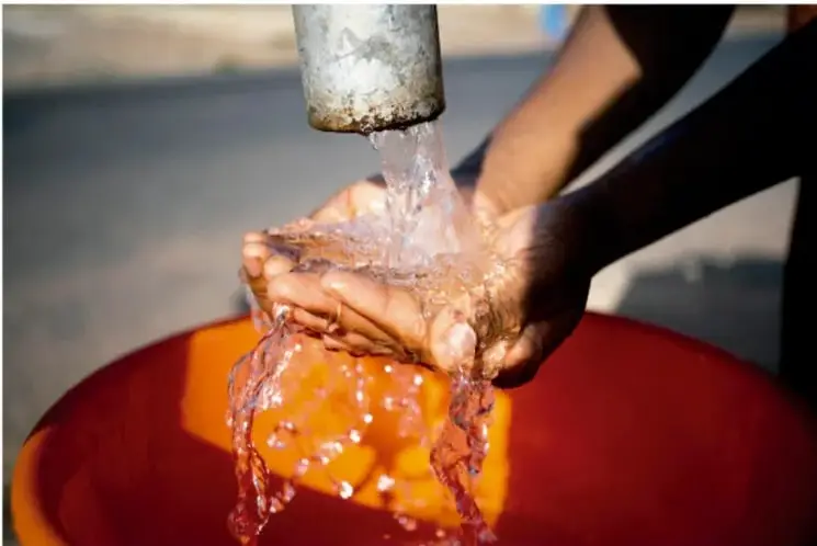 Fotografía de unas manos recibiendo agua que cae de un grifo, sobre un recipiente rojo, en un entorno exterior.