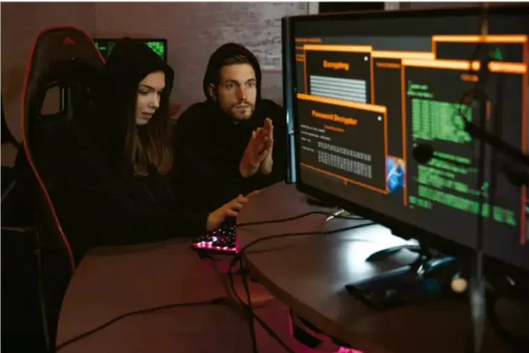 Fotografía de dos personas jóvenes sentadas frente a una computadora que muestra ventanas con código y herramientas de cifrado y descifrado de contraseñas, en un ambiente oscuro iluminado por la pantalla, ilustrando el trabajo en ciberseguridad.