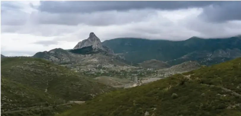 Fotografía de un paisaje montañoso con una peña prominente, bosque y un valle habitado bajo cielo nublado.