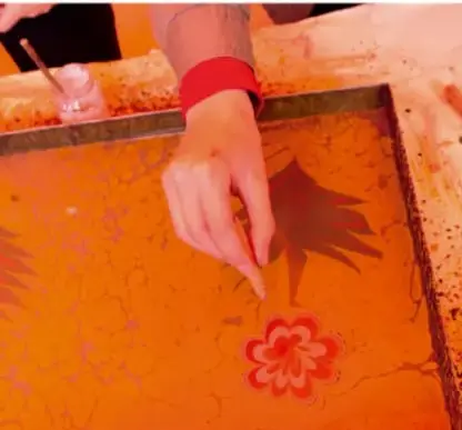 Mano deslizando pintura naranja con una herramienta sobre una malla de serigrafía con un diseño de flor, ilustrando el procedimiento manual de la técnica.