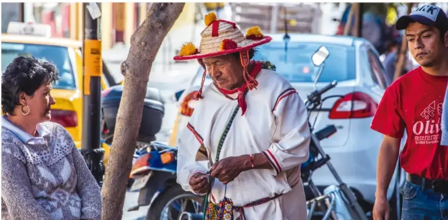 Fotografía de una calle urbana en México: una mujer mayor conversa con un hombre que usa sombrero tradicional artesanal con flores y traje blanco bordado, mientras un joven con camiseta roja y gorra pasa cerca; al fondo hay autos y motocicletas, ilustrando la convivencia cotidiana de culturas diversas.