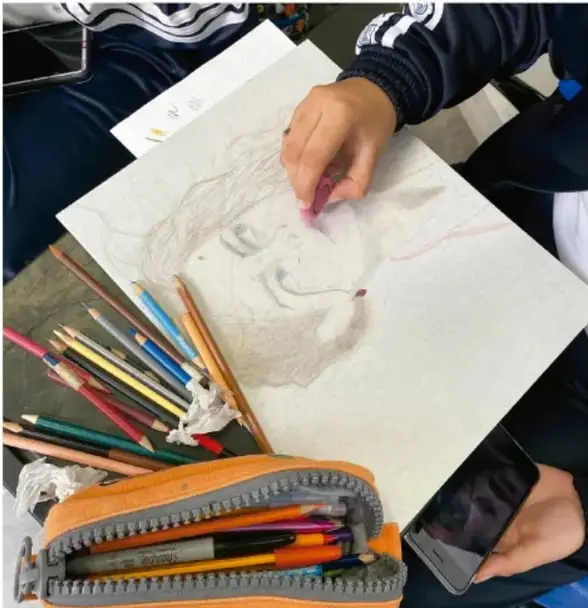 Foto de estudiantes trabajando en el mural: mano dibujando con lápices de colores, estuche con lápices, hoja con boceto de rostro y un teléfono celular; crédito: Gimenna Schiaffini Rosales, PUEBLA.
