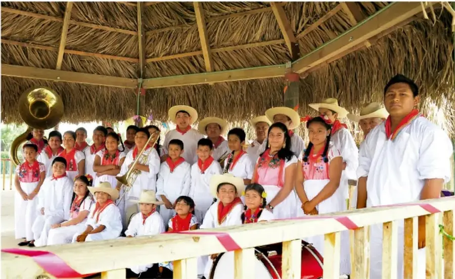 Grupo de personas, adultos y niños, con vestimenta tradicional, algunos con instrumentos musicales, bajo un techo de palapa.