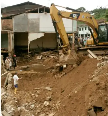 Maquinaria excavadora CAT trabajando entre escombros y lodo, con personas alrededor.