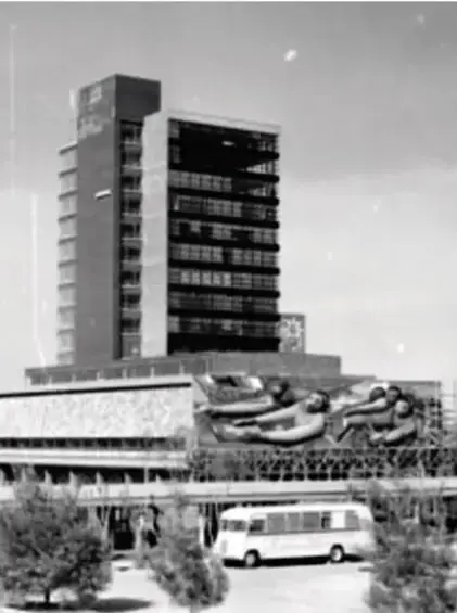 Fotografía en blanco y negro de un edificio moderno de varios pisos con una construcción al frente y un autobús estacionado.