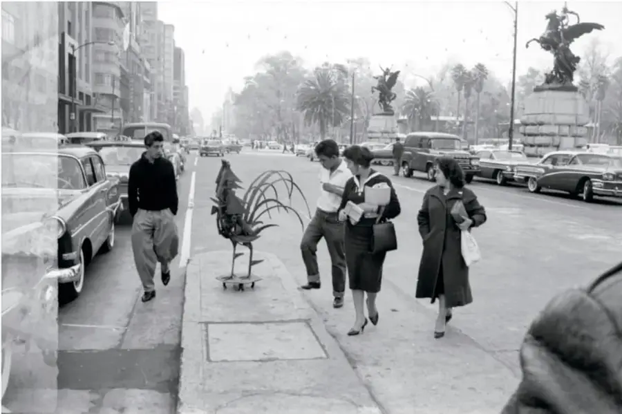 Fotografía en blanco y negro: varias personas en la banqueta de una avenida amplía se detienen a observar un gallo de hierro forjado colocado sobre la acera; hay autos antiguos estacionados y, al fondo, monumentos y esculturas de la Ciudad de México.