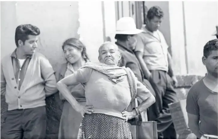 Grupo de personas en la calle: un joven de suéter, una señora con rebozo y las manos en la cintura, y varios vecinos más. Fotografía en blanco y negro titulada 'La celestina, de la serie Candelaria de los Patos, 1963'.