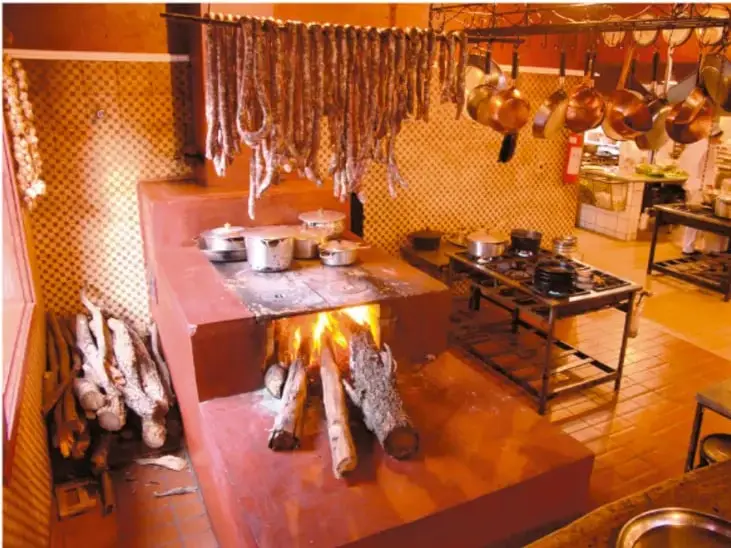 Fotografía de una estufa ahorradora de leña encendida, con troncos de madera y utensilios de cocina alrededor.