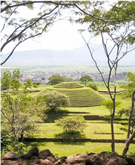 Vista panorámica de una pirámide circular escalonada entre árboles, con el valle y el pueblo de Teuchitlán al fondo.