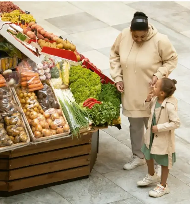 Foto de una madre y una niña frente a un puesto de verduras y frutas del mercado; se muestran zanahorias, papas, brócoli y otros vegetales.