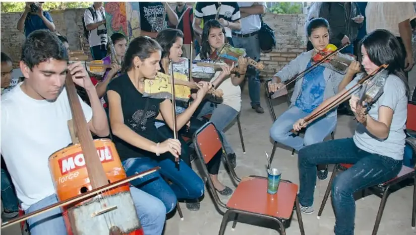 Fotografía de un grupo de estudiantes sentados en sillas, tocando instrumentos de cuerda construidos con materiales reciclados (cajas, tubos y cuerdas), acompañados por adultos. Representa la presentación musical del proyecto.