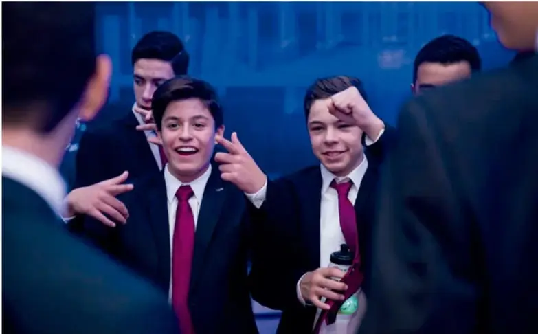 Fotografía de un grupo de estudiantes adolescentes con traje formal, corbata roja y corbata verde, conversando yriendo en un evento; uno de ellos sostiene un termo. Ilustra el uso de las expresiones en una conversación real.