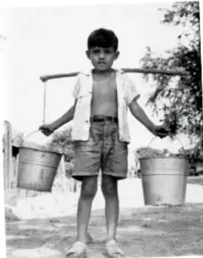 Fotografía en blanco y negro de un niño descalzo que camina cargando una cubeta colgada de cada brazo con un palo al hombro, ilustrando el acarreo de agua.