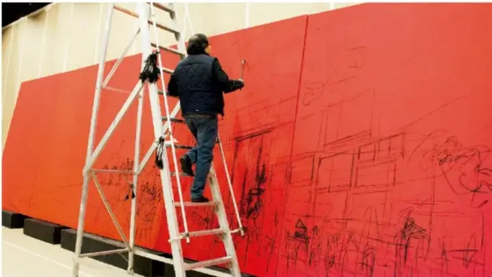 Persona subida a una escalera pintando de rojo un muro grande con un boceto a lápiz.