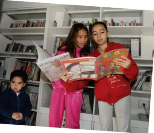 Dos niñas y un niño hojean libros y revistas en la biblioteca escolar.