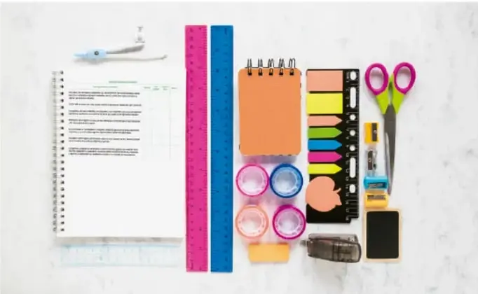 Fotografía de materiales escolares sobre fondo blanco: agenda abierta, reglas, cuaderno naranja, marcadores de colores, tijeras, clips, borrador, pegamento y notas adhesivas.
